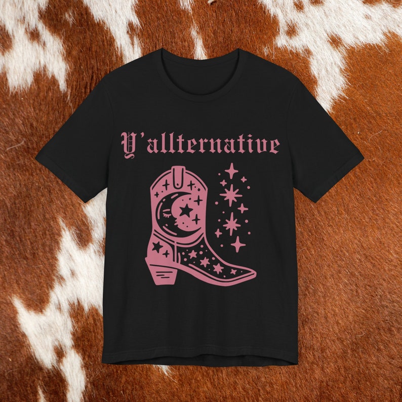 Puede incluir: Camiseta negra con un gr&aacute;fico rosa de una bota vaquera con estrellas y una luna creciente. El texto "Y'allternative" est&aacute; impreso sobre la bota.
