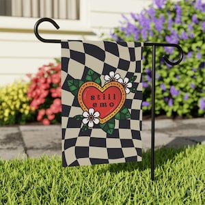 May include: A garden flag with a black and white checkered pattern. A red heart with a yellow outline and the words "still emo" is in the center of the flag. The heart is surrounded by green leaves and white flowers.