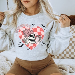 Peut inclure: Sweat-shirt gris avec un motif de c&oelig;ur &agrave; carreaux rose et blanc. Le c&oelig;ur repr&eacute;sente un squelette portant un bonnet rose et tenant une tasse de caf&eacute;. Le texte "Spooky Moms Club" est &eacute;crit au-dessus du c&oelig;ur.