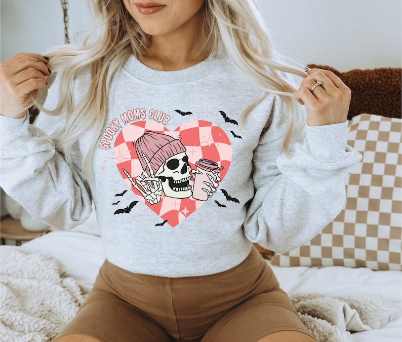 Peut inclure: Sweat-shirt gris avec un motif de c&oelig;ur &agrave; carreaux rose et blanc. Le c&oelig;ur repr&eacute;sente un squelette portant un bonnet rose et tenant une tasse de caf&eacute;. Le texte "Spooky Moms Club" est &eacute;crit au-dessus du c&oelig;ur.