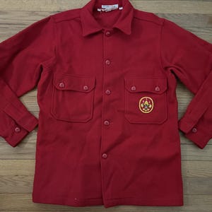 May include: A red button-up shirt with long sleeves, two chest pockets, and a collar. The shirt features a yellow and black embroidered patch on the left pocket. The shirt is made of a thick, possibly wool, material.