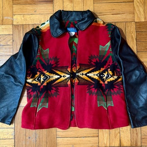 May include: A red jacket with black leather sleeves and collar. The front of the jacket features a geometric pattern in yellow, green, and black. The jacket is open, revealing a zipper closure.