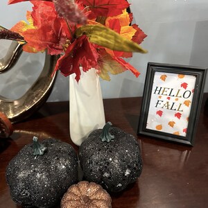 Könnte beinhalten: Eine herbstliche Dekoration mit einer weißen Vase mit Herbstblättern und dekorativen Kürbissen. Zwei schwarze Glitzerkürbisse und ein kleinerer Bronzekürbis sind zusammen mit einem gerahmten Schild mit der Aufschrift "HELLO FALL" angeordnet.