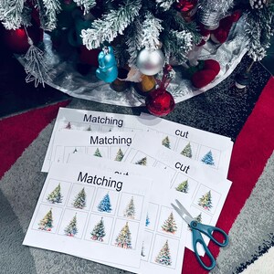 May include: A set of printable Christmas tree matching and cutting activity sheets. The sheets feature colorful illustrations of Christmas trees with different decorations. The sheets are printed on white paper and include the words "Matching" and "Cut". A pair of blue scissors is also included in the image.