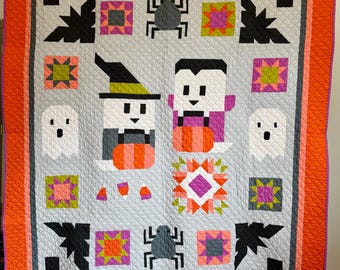 Könnte beinhalten: Eine Halloween-Steppdecke mit Patchwork-Design, die verschiedene Figuren und Symbole zeigt. Die Steppdecke ist aus verschiedenen Stoffen in Orange, Pink, Grün und Grau gefertigt. Zu den Figuren gehören eine Hexe, ein Vampir und Geister. Zu den Symbolen gehören Spinnen, Mais und Sterne.
