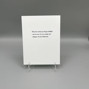 May include: A white rectangular greeting card featuring black printed text that reads: When the witches go riding, and black cats are seen, the moon laughs and whispers, 'tis near Halloween. The card is supported by a clear acrylic stand.