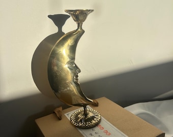 May include: A brass candle holder in the shape of a crescent moon with a carved face. The candle holder has a floral base and a small cup for the candle. The moon sits on a cardboard box with text on it.