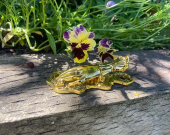 Puede incluir: Un llamador de puerta decorativo dorado con forma de mano que sostiene un ramo de flores. El llamador descansa sobre una superficie de madera envejecida, con pensamientos amarillos y morados en el fondo.