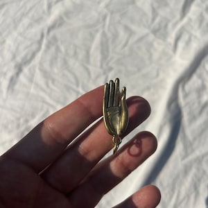 Peut inclure: Un charme en métal doré en forme de main avec un anneau attaché au poignet. Le charme est suspendu à une chaîne.
