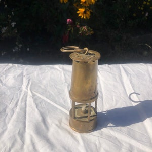 Op de afbeelding: Een messing mijnwerkerslamp met een haak aan de bovenkant en een draadkooi rond het glas. De lamp staat op een wit oppervlak.