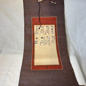 Peut inclure: Un parchemin japonais vintage avec calligraphie à l'encre noire sur papier vieilli. Le parchemin présente un tissu à motifs violet foncé avec une bordure rouge autour du texte central. Des tiges en laiton sont en bas.