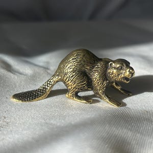 Könnte beinhalten: Kleine, detaillierte Biberfigur aus Messing. Die Skulptur weist eine strukturierte Fellstruktur, einen flachen Schwanz und eine dynamische Pose auf. Der Biber hat eine warme, goldene Farbe und steht vor einem weißen Hintergrund.