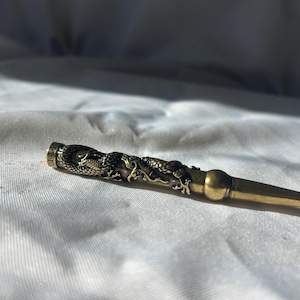 May include: A brass-coloured metal pipe with a dragon design. The pipe has a detailed dragon wrapped around the body and a tapered end. The pipe is lying on a white surface.