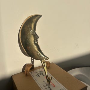 Pode incluir: Um gancho de meia-lua cor de latão com um rosto, montado em um pequeno suporte. O gancho é preso a uma caixa de papelão com um rótulo com caracteres e ilustrações asiáticas. A lua tem um rosto detalhado.