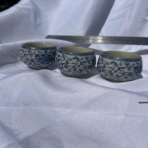 Pode incluir: Três pequenas taças de cerâmica redondas com um padrão floral azul e branco. As taças estão posicionadas em frente a uma régua metálica com medidas em centímetros, indicando que têm um diâmetro de aproximadamente 6,5 cm.