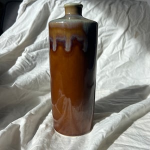 May include: A brown ceramic vase with a light brown glaze and a white drip glaze. The vase has a cylindrical shape and a rounded bottom.