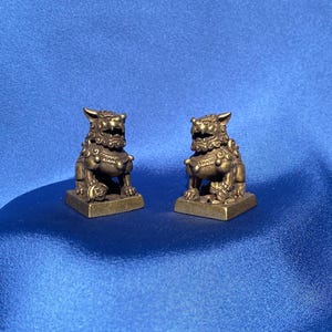 May include: Two small gold-colored lion statues on a blue background. The statues are facing each other and have intricate details.