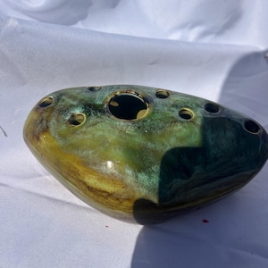 May include: A ceramic flower frog with a smooth, glossy finish in shades of deep forest green, teal, and earthy mustard yellow. The object has an irregular, rounded shape with one large central opening and several smaller holes for stems.