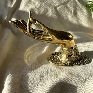 May include: A gold-colored metal hand sculpture with an ornate base. The hand is in a gesture of giving or receiving, with the fingers curled inward.