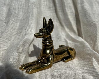 May include: A metallic gold-toned figurine of a jackal in a reclining position, featuring pointed ears, a smooth neck collar, and a sleek, elongated body. The sculpture has a polished finish with subtle dark shading in the crevices.