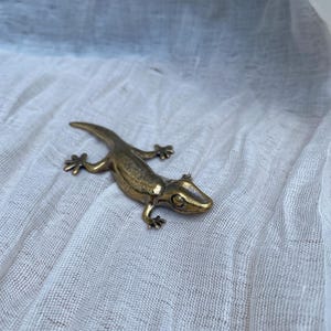 May include: A small, metallic bronze-colored lizard figurine with a textured surface, depicted in a crawling position with four legs extended and a long, curved tail. The lizard features detailed eyes and toes, showcasing a realistic anatomical shape.