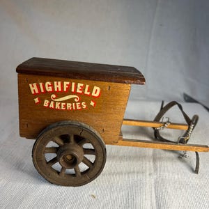 Può includere: Un carrello in legno in miniatura con finitura marrone, con una singola ruota e una scatola con la scritta "HIGHFIELD BAKERIES" in lettere rosse e bianche. Il carrello ha un tetto marrone scuro ed è progettato con un manico.