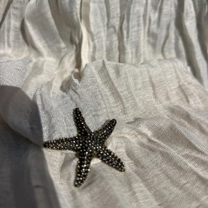 Op de afbeelding: Een donkere, getextureerde broche in de vorm van een zeester met een dicht oppervlak van kleine, reflecterende kralen of kristallen, rustend tegen een achtergrond van gebroken witte, gekreukelde, semi-transparante stof met verticale plooien.
