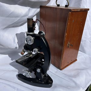 May include: A vintage black microscope with silver controls and a wooden case. The microscope features a curved arm and a stage for slides. The wooden case is rectangular with a metal handle. The items are on a white surface.