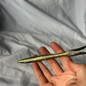 May include: A gold-colored, fish-shaped serving utensil. The utensil has a detailed scale pattern and a forked tail. The tongs are held open, ready for use. The background is a plain, white fabric.