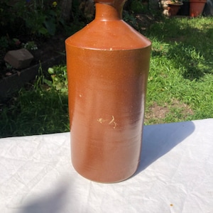 May include: A brown ceramic bottle with a wide mouth and a slightly flared lip. The bottle has a rustic, vintage look.