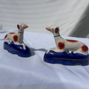 May include: Two ceramic dog figurines. The dogs are white with red spots and a black collar. They are perched on a blue base with white wave details. The figurines are likely decorative items.