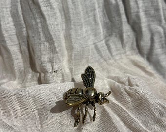 Peut inclure: Une broche en métal en forme d'abeille avec une finition en laiton antique, présentant des ailes détaillées et un corps segmenté. Le design de l'insecte comprend de petites pattes et des antennes, avec une surface texturée soulignant le travail.