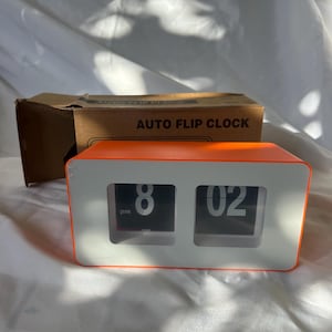 May include: An orange and white auto flip clock displaying 8:02 pm. The clock has a rectangular shape with a white face and orange trim. The cardboard box behind the clock reads "AUTO FLIP CLOCK".