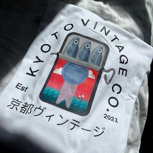 May include: White t-shirt with a vintage logo featuring a red, white and blue can of fish with three fish inside. The text on the shirt reads "Kyoto Vintage Co. Est 2021" and "京都 ヴィンテージ".