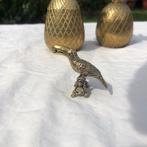 Puede incluir: Una figurita de pájaro tucán de oro con un diseño detallado. El tucán está posado en una rama de árbol con el pico abierto. La figurita está sentada sobre una superficie blanca.
