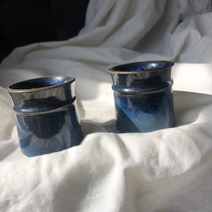 May include: Two blue ceramic mugs with a dark glaze. The mugs are sitting on a white fabric surface.