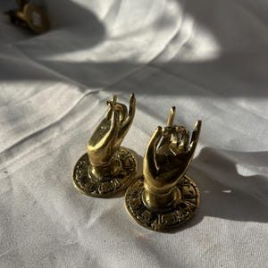 Könnte beinhalten: Zwei goldfarbene, handförmige Haken aus Metall mit filigranen Details. Die Hände sind in einer Geste des Gebens oder Empfangens. Die Haken sind auf runden Sockeln mit dekorativen Mustern montiert.