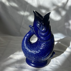 May include: A cobalt blue ceramic fish-shaped jug. The jug has a textured scale pattern and a wide open mouth. The handle is formed by the fish's tail. The base is round and the jug is approximately 18 cm tall.