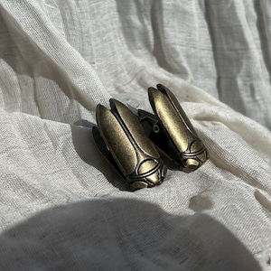 Puede incluir: Un par de clips de metal en tono latón antiguo con forma de escarabajos. Cada clip presenta un cuerpo liso y alargado con detalles segmentados y un diseño de cabeza curva, mostrando un acabado metálico con sutiles reflejos oscuros.