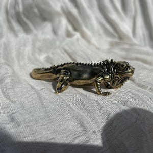 May include: A small, metallic, brass-coloured lizard figurine with a textured back, detailed scales, and a prominent crest along its spine. The lizard is depicted in a crawling position with its four legs extended and its head turned slightly to the side.