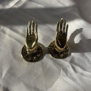 以下が含まれることがあります： 複雑なディテールが施された、金色の金属製のハンドシェイプのドアノブ2つ。ノブは、装飾的なデザインが施された丸いベースに取り付けられています。