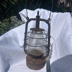 May include: A weathered, rusted metal lantern with a clear glass globe protected by a wire cage. The lantern features a top handle and a circular base. A metal ruler is positioned vertically next to the base, showing measurements in centimetres.