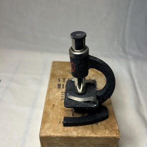 Puede incluir: Un microscopio negro vintage con un ocular y una platina plateados, apoyado sobre una caja de cartón. La caja tiene texto impreso, incluyendo "STAMP MAGNIFIERS". El microscopio presenta un brazo curvo y un emblema rojo circular.