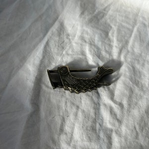May include: A silver-toned metal fish-shaped padlock with intricate details. The padlock is closed and has a small keyhole on the side.