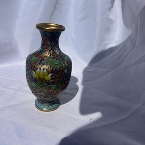 May include: A small metal vase featuring intricate cloisonné enamel work with a vibrant floral pattern in shades of blue, green, red, and yellow. The vase has a narrow neck, a flared rim, and a circular base, all finished with gold-toned metal edges.
