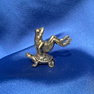 Könnte beinhalten: Eine goldfarbene Froschfigur, die auf einer Schildkrötenfigur sitzt. Der Frosch sitzt aufrecht mit ausgestreckten Beinen, während die Schildkröte mit Kopf und Beinen sichtbar nach vorne geht.