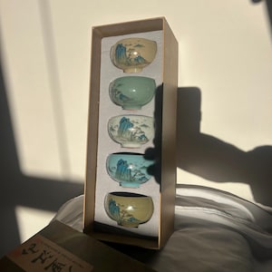 Könnte beinhalten: Ein Set aus vier kleinen Keramik-Teetassen in einer rechteckigen Box. Jede Tasse zeigt eine Landschaftsmalerei in Blau-, Grün- und Gelbtönen. Die Tassen sind in einer beigefarbenen Box präsentiert. Die Box ist teilweise geöffnet und zeigt den Inhalt.