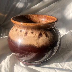 May include: A brown and tan glazed ceramic planter with a rounded shape and a wide rim. The planter has a brown base.