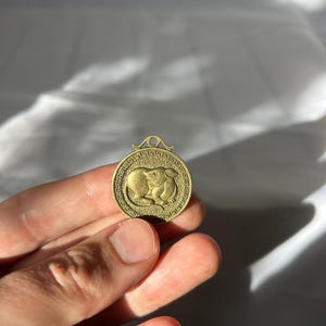 Könnte beinhalten: Ein runder, goldfarbener Anhänger mit einem detaillierten Relief einer Ratte innerhalb eines dekorativen Randes. Der Anhänger hat oben eine kleine Schlaufe zum Aufhängen. Das Design ist wahrscheinlich von chinesischen Tierkreiszeichen inspiriert.