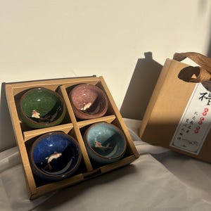 May include: A set of four small ceramic bowls in a wooden box. Each bowl is a different color: green, pink, blue, and teal. Each bowl features a painted fish. A gift box with text is next to the bowls.